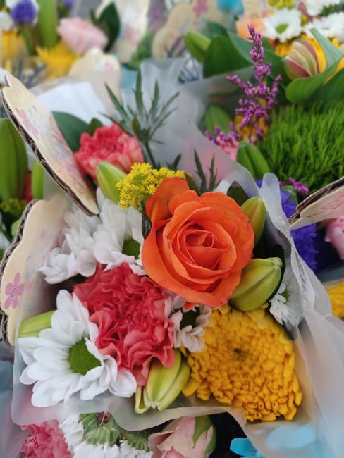 A vibrant floral arrangement featuring a prominent peach-orange rose at the center, surrounded by pink carnations, white daisies with green centers, yellow chrysanthemums, and sprigs of purple and yel