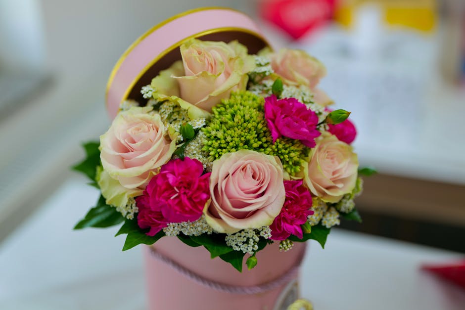 A round floral arrangement in a pastel pink cylindrical gift box, featuring soft pink roses with delicate, velvety petals, vibrant magenta carnations with ruffled blooms, clusters of small white and g
