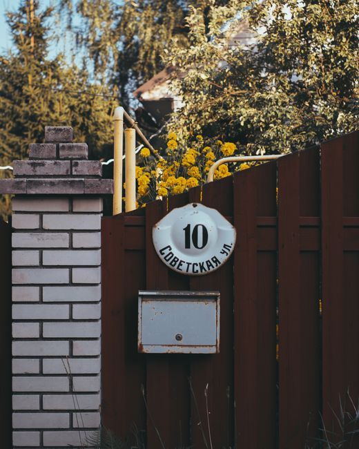 A close-up of a red wooden fence with a white circular street sign displaying the number 10 and Cyrillic text beneath, mounted on the fence. Behind the fence, there are yellow flowers in full bloom an