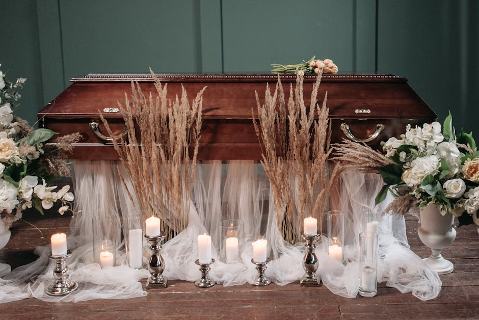A wooden funeral casket resting on a wooden floor, decorated with tall arrangements of beige pampas grass and two large bouquets of white and cream flowers including roses and orchids in white vases o