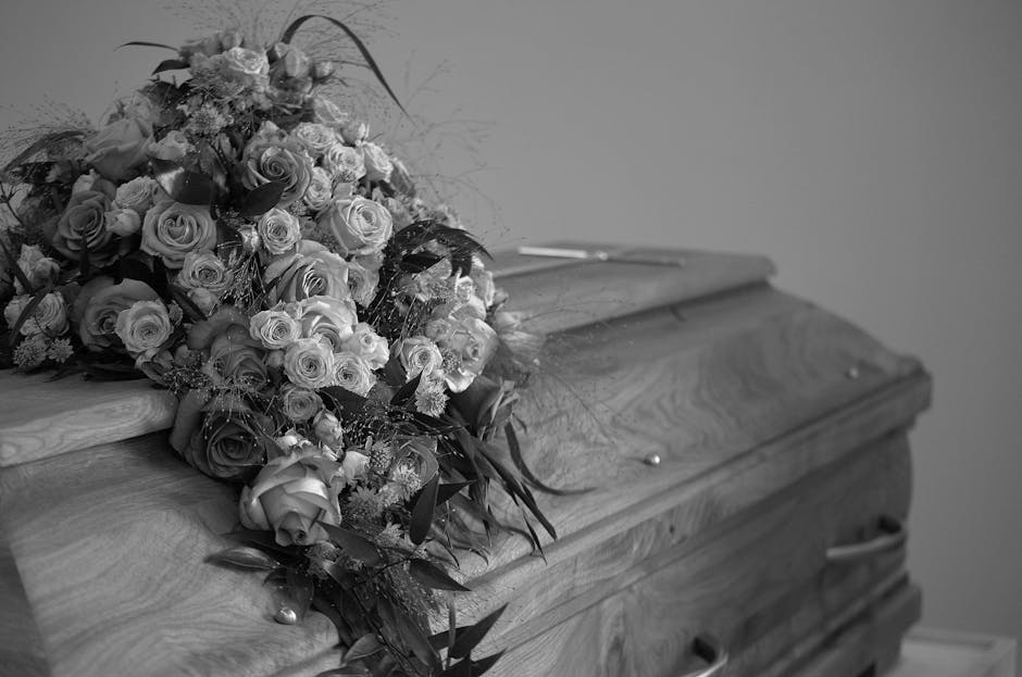 A large, elegant funeral spray resting on a polished wooden coffin, composed of numerous fresh roses in varying shades of light and dark, with some roses partially open and others in full bloom. The a